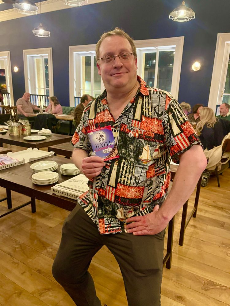 The image shows Nick Steel the director of Bath Comedy Festival showing off the new festival programme. He is wearing a bright shirt sat on a table.