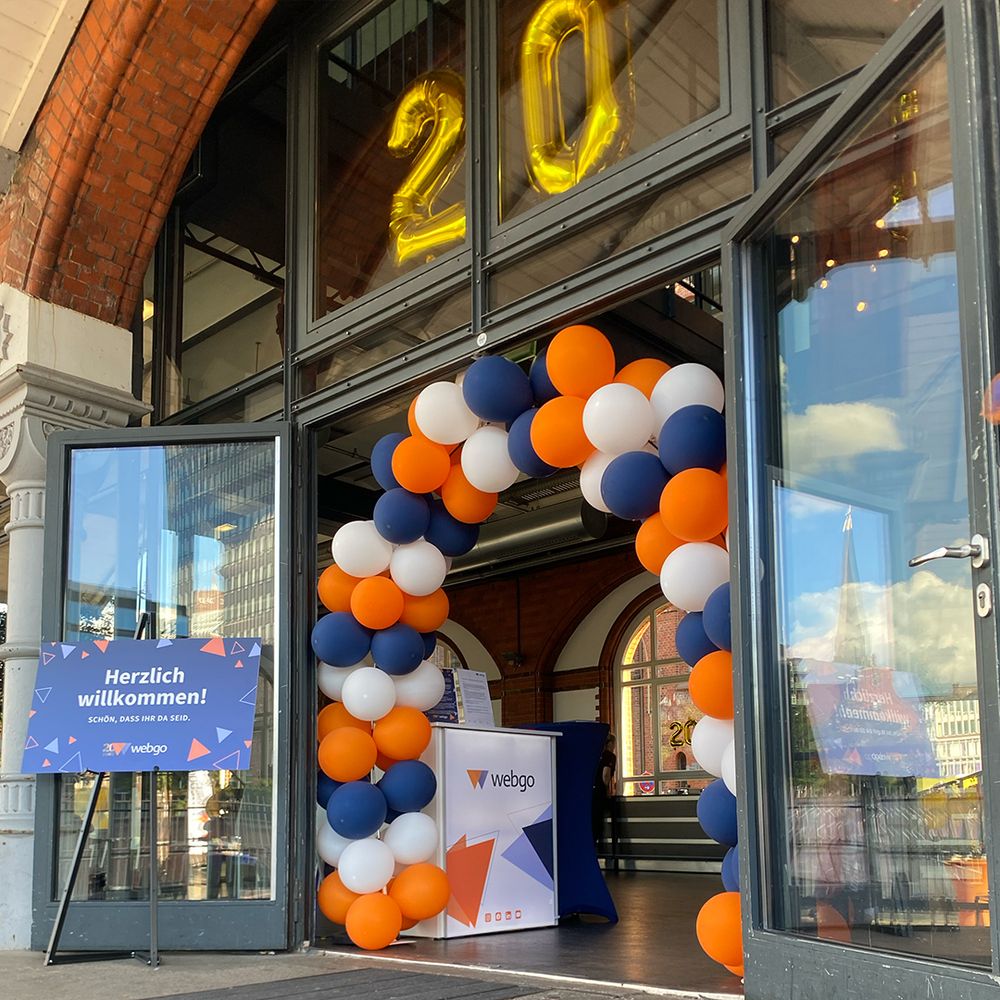 Foto vom Eingang der Event-Location zur 20-jährigen Jubiläumsfeier der webgo GmbH. Ein Ballonbogen mit orangefarbenen, weißen und blauen Ballons, sowie ein Willkommensschild und Helium-Ballons mit der 20.