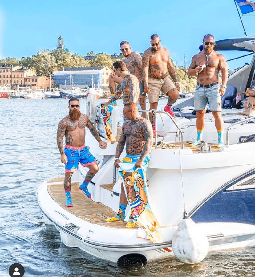 Jan Emanuel och sex tatuerade gymkillar poserar på en båt iförda shorts och färgglada strumpor. 
