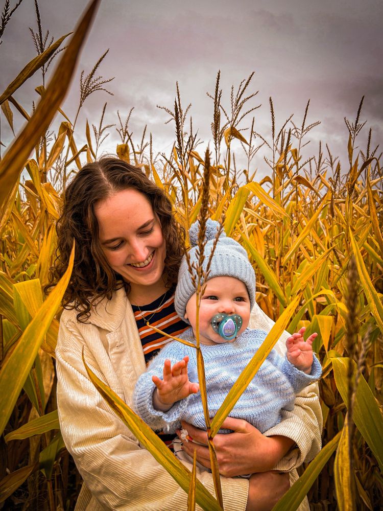A woman holds a 10 month old baby wearing blue knit clothes in a corn field, both looking happy