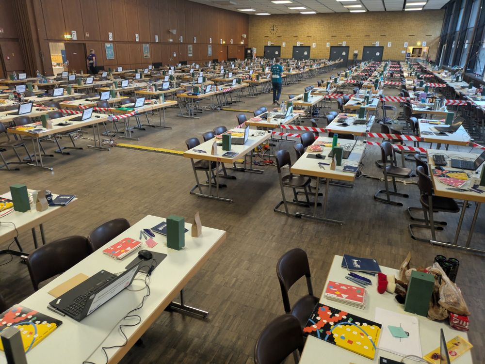 Picture of the contest hall (transformed mensa) with many tables in a grid-like setting. Each table has a laptop and 3 chairs, and notebooks and an envelope with problem statements.