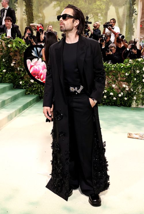 Sebastian Stan at the 2024 met gala. He’s wearing an all black outfit with black sunglasses. His hair his slick back straight. 