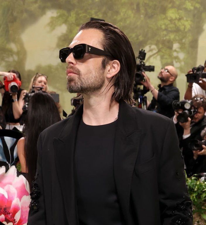 Sebastian Stan at the 2024 met gala. He’s wearing an all black outfit with black sunglasses. His hair his slick back straight. 