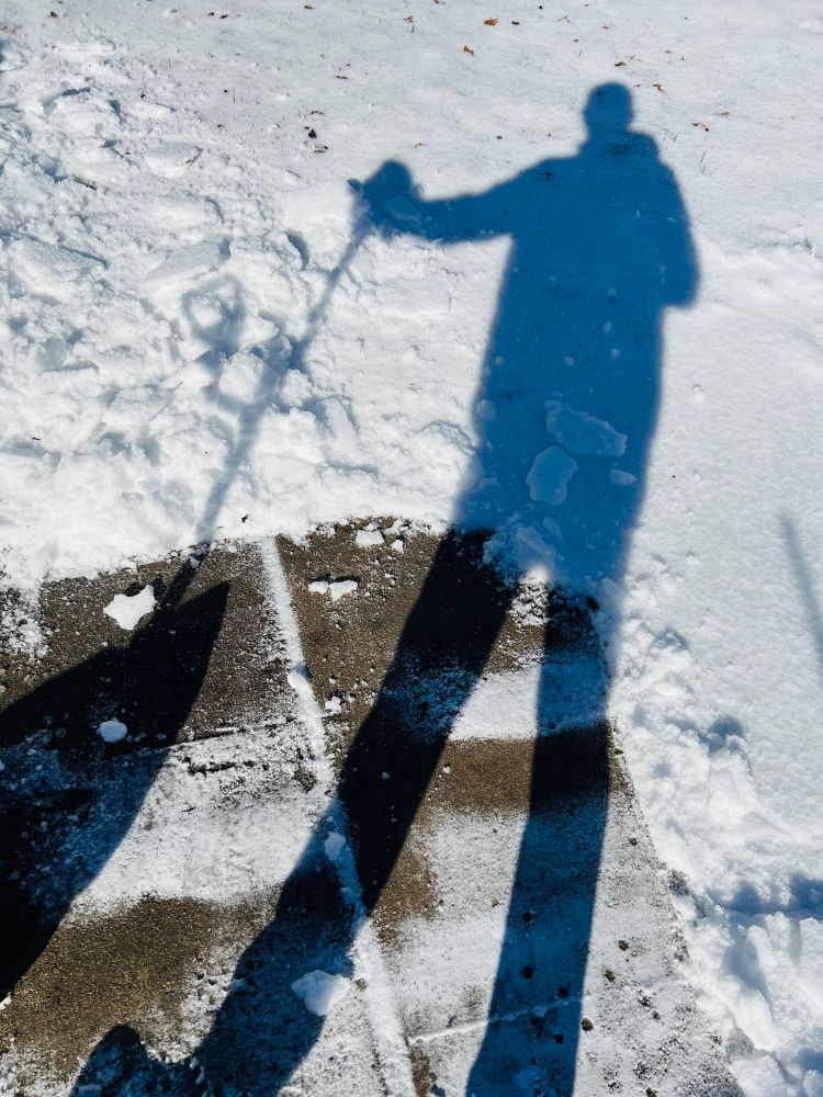 Shadow of me standing with a shovel cast upon the snow with a freshly cleared sidewalk. 