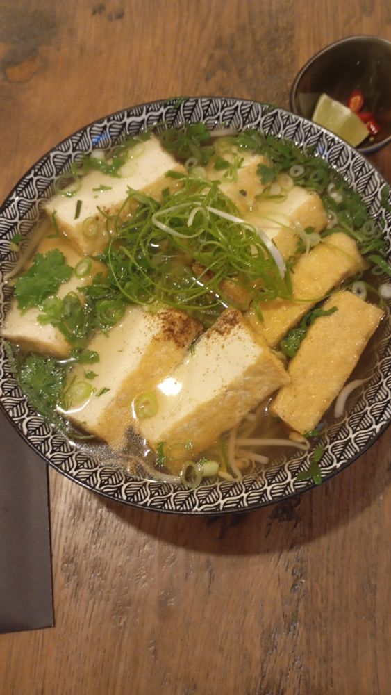 a bowl of pho topped with thinly sliced spring onions and slices of fried tofu. served with a slice of lime and a few slices of hot peppers
