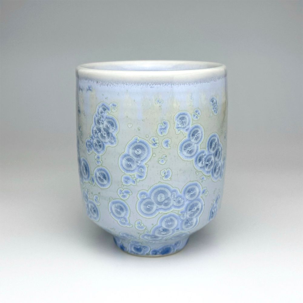 A ceramic cup glazed with a light blue zinc crystalline glaze. The background of the glaze is mix of creamy warm colors and periwinkle blue, while the crystals are alternating rings of varying shades of light blue. 