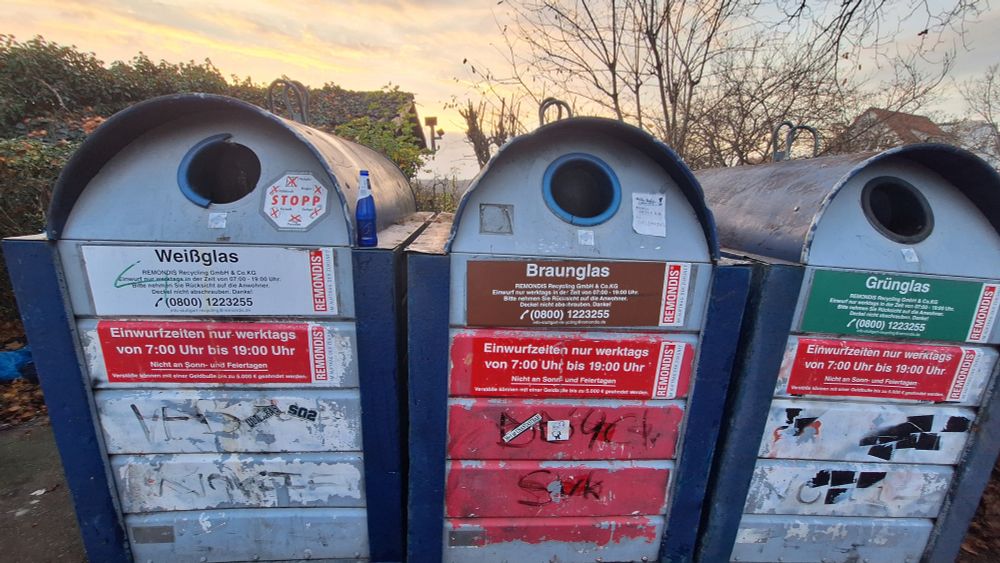 Drei Altglascontainer für weiß-, braun- und Grünglas stehen nebeneinander. auf einem davon ist eine blaue Glasflasche abgestellt.