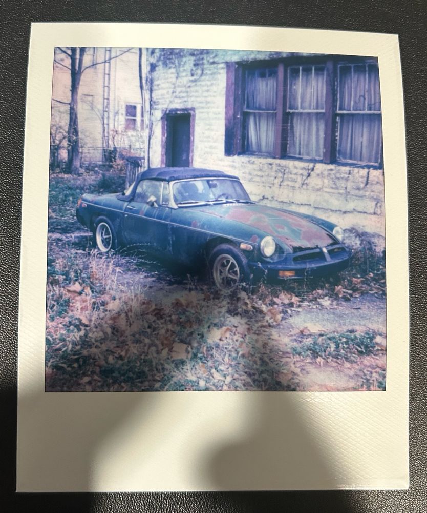 A Polaroid picture of a rusted MG care in front of a run down building. There are leaves, long grass, and weeds all around.