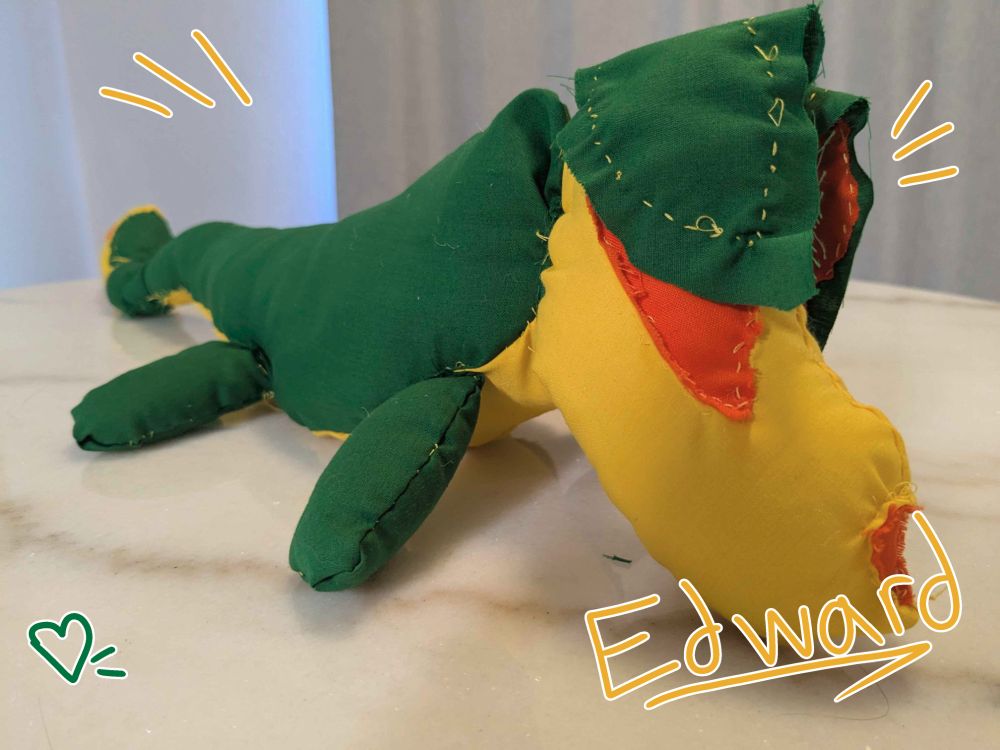 A plushie made of orange, yellow, and green fabric, with yellow thread, of Edward the fruity fox, on a white table