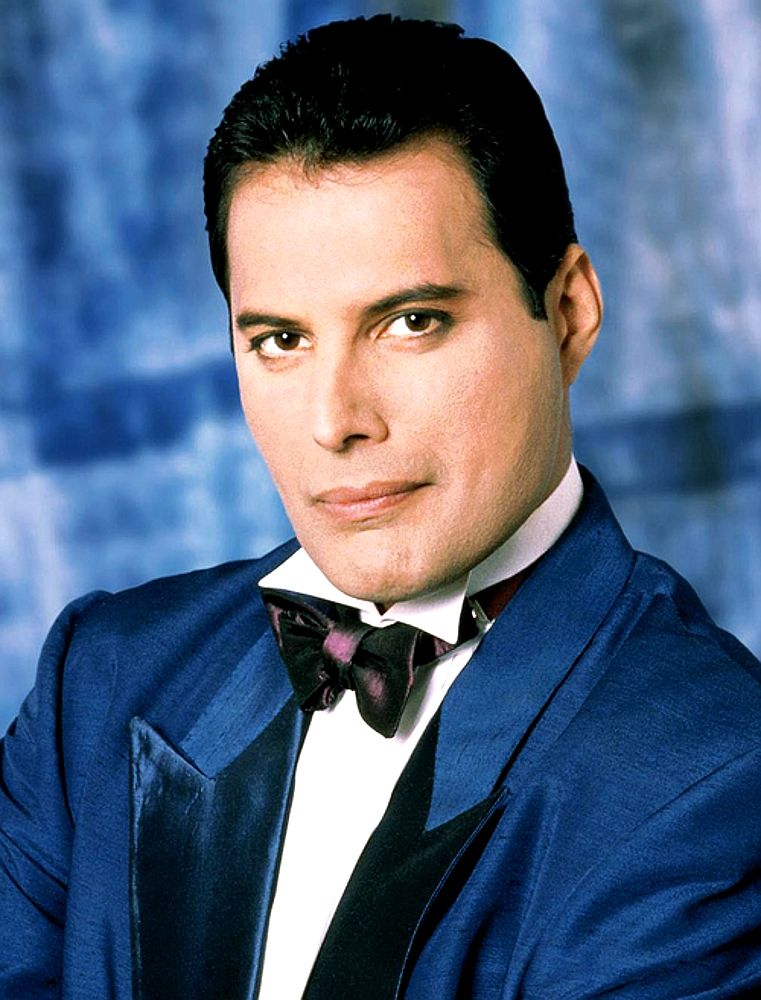 Freddie Mercury in 1988. He has short hair and no moustache. He is wearing  a blue tuxedo, white shirt and purple velvet bow tie