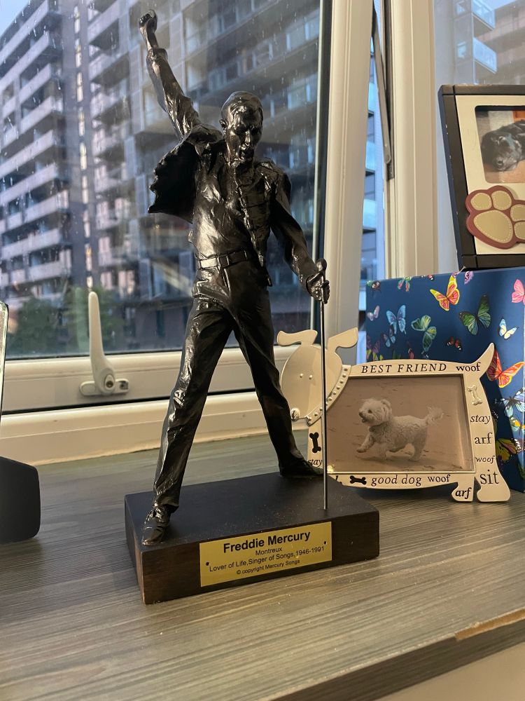 Metal replica of Freddie Mercury statue on a windowsill. He is in his famous pose with one arm in the air and his mic stand to the ground. The plaque on the wooden base says ‘Freddie Mercury. Montreux. Lover of life, singer of songs 1946-1991.’ There is a window in the background showing tower blocks in the rain, and a framed photograph of fluffy white dog beside the statue.