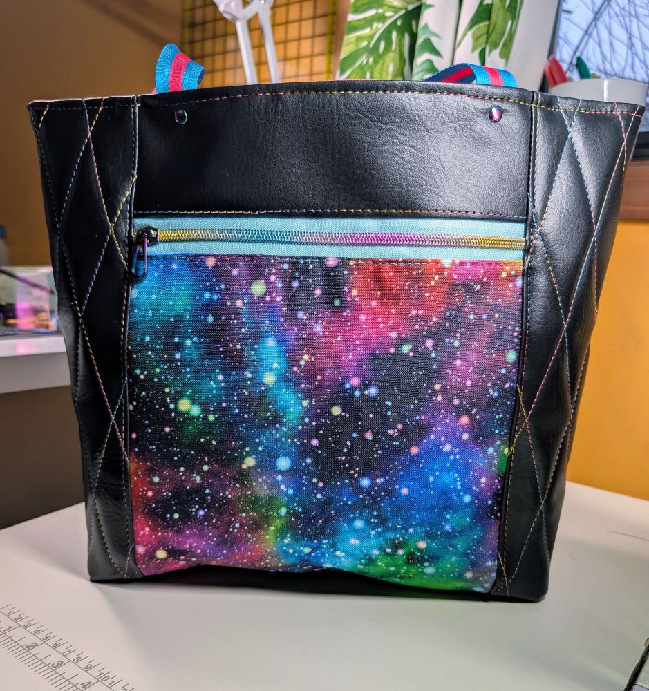 A picture of a tote bag made of black faux leather, quilted in a diamond pattern with rainbow variegated thread. The center front panel of the tote is a bright print space print. 