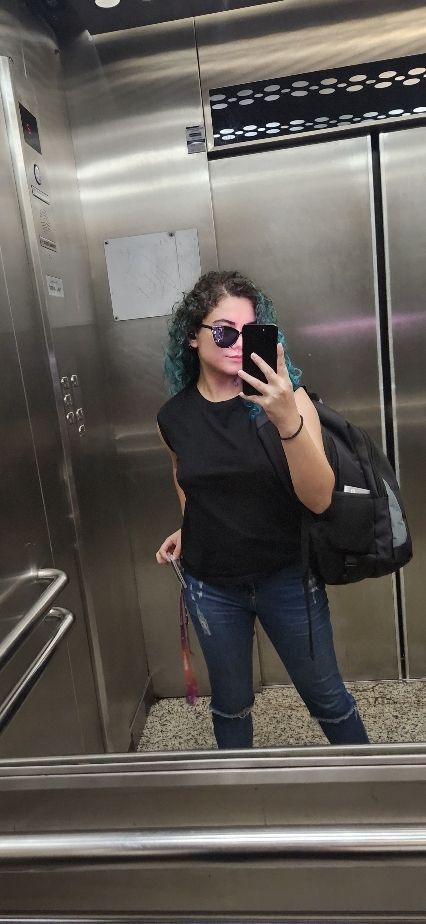 Mulher branca, cabelo cacheado azul, vestindo uma regata preta e jeans claro, usando óculos escuro e com uma mochila nas costas. Foto no espelho de um elevador usando o celular. 