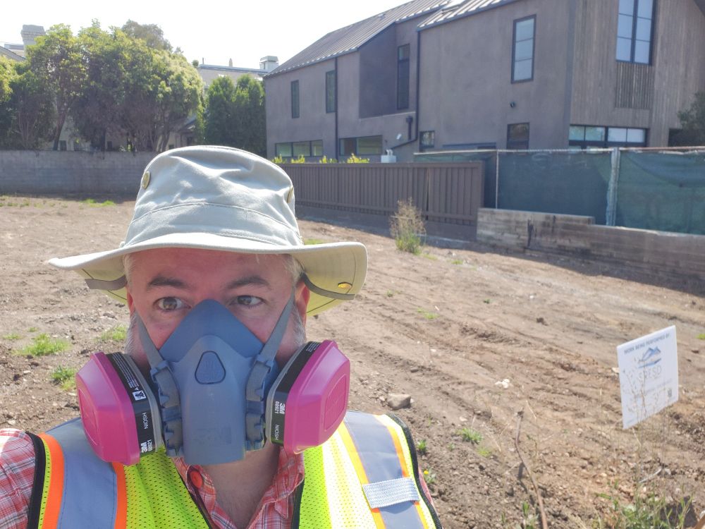 A man in a hat and high vis vest wearing a P100 respirator. Behind is an empty house lot, with a house standing on the lot next door.