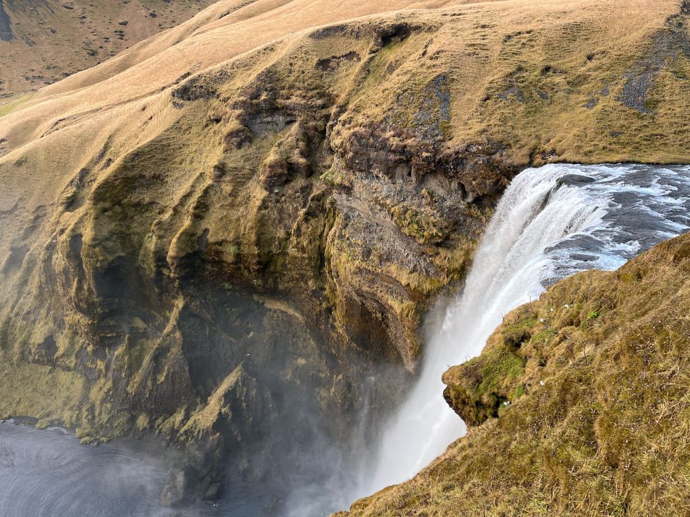 Skógafoss waterfall, Iceland. The grass and moss-covered hill side ends abruptly with a sheer drop which the shallow rocky river falls over.