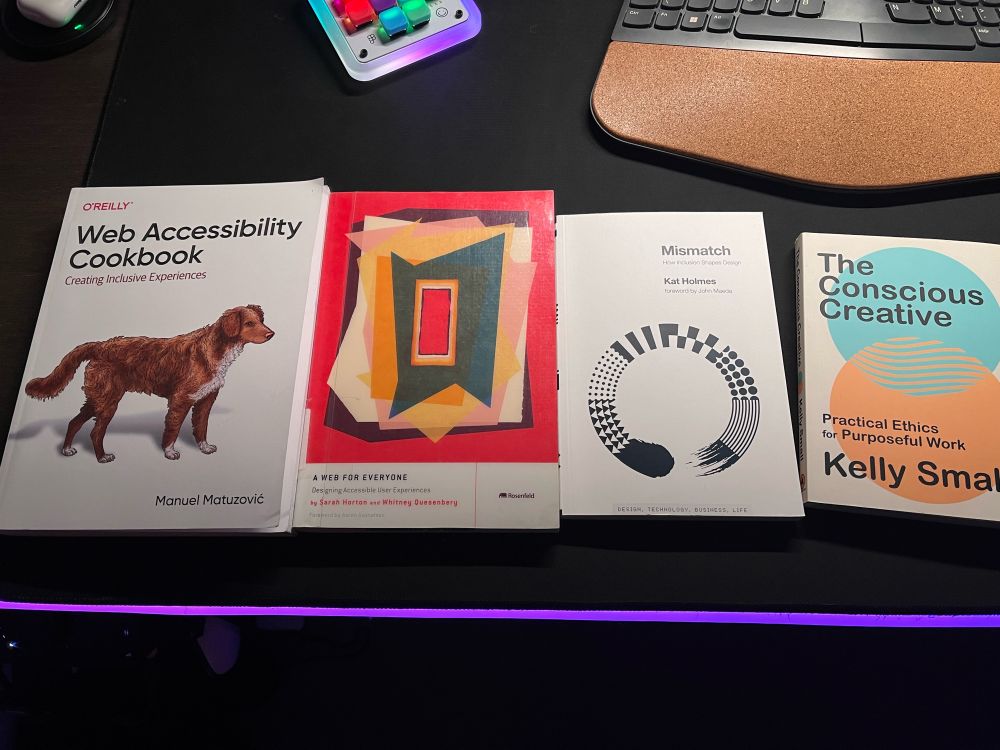 Four books laid out on a black desk lined with lights; Web Accessibility Cookbook by Manuel Matuzović, A Web For Everyone by Sarah Horton and Whitney Quesenbery, Mismatch by Kat Holmes, and The Conscious Creative by Kelly Small.