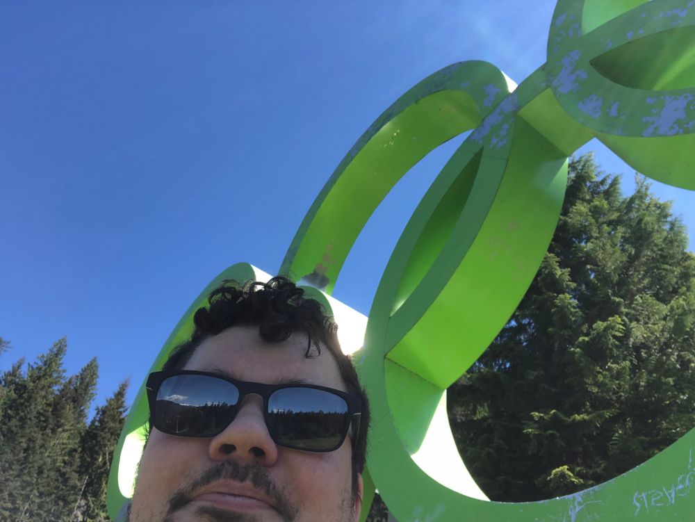 Lucas (white man in sunglasses and beard, sweaty) standing right in front of bright green Olympic rings