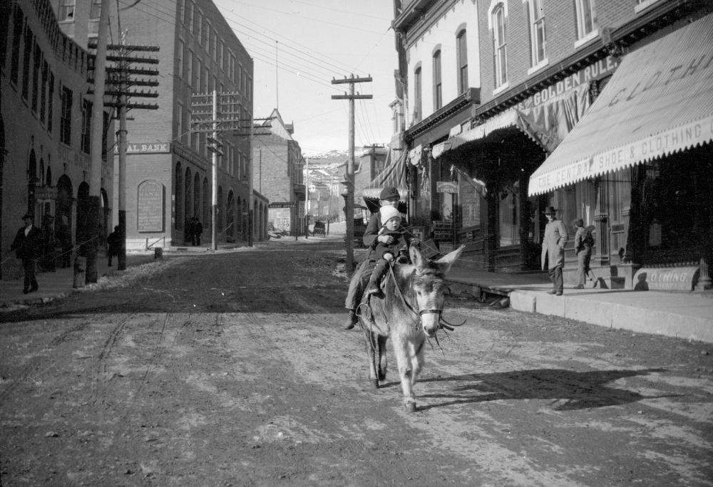 Central City | circa 1900 | A boy and toddler ride a burro down Eureka Street while a man watches from sidewalk. Signs visible include "The Golden Rule" and "First National Bank."
