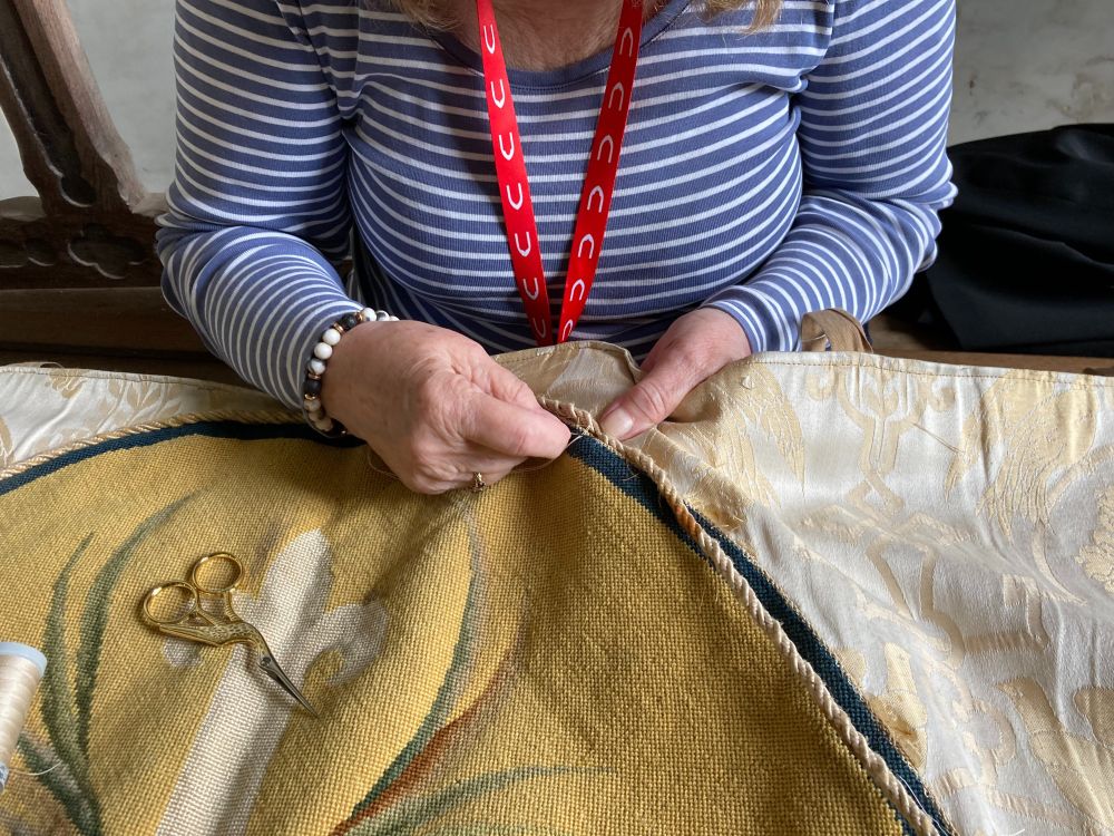 Close up of Wendy's hands at work, sewing the cord into place. A golden pair of thread scissors in the shape of a bird rests on the altar frontal.