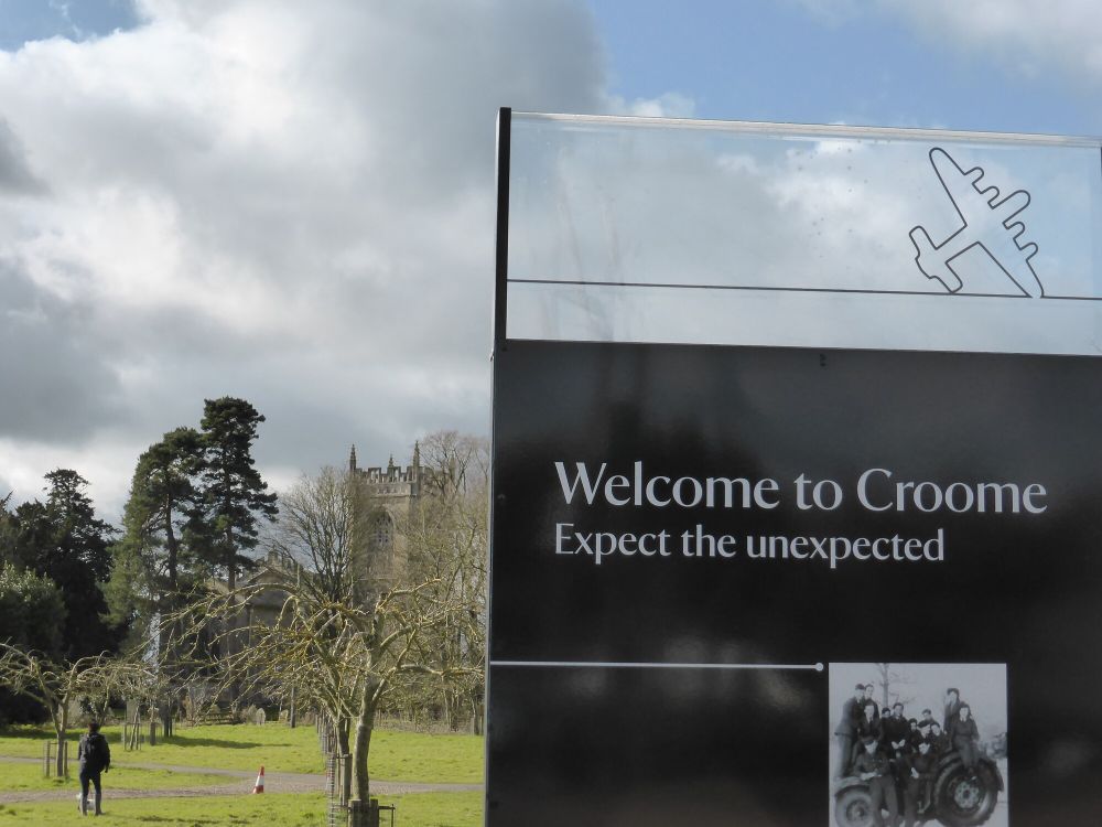 Photograph of a sign at Croome estate with large silver text on black ground reading 'Welcome to Croome' and smaller silver text on black ground below this reading 'Expect the unexpected' underneath the text is a black and white photograph, above the text is a black line drawing of a WWII plane on silver ground. Behind the sign in the distance we can see St. Mary Magdalene church behind trees. (c) Tom Jones