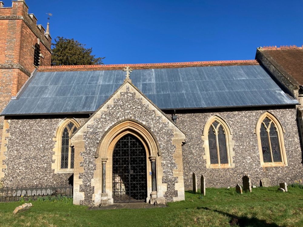 Full view of St. Bartholomew's, Lower Basildon, a historich church with flint covered porch and nave, three tall nave windows, a crenellated red brick tower and a corrugated iron roof.