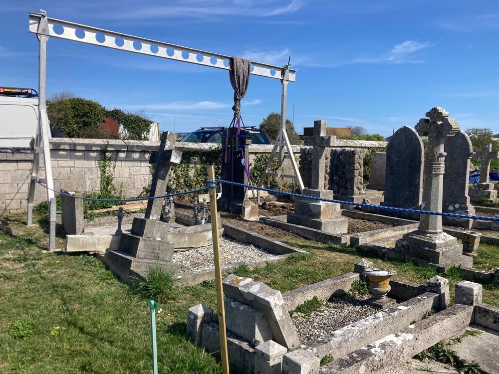 A gravestone being suspended from a metal beam, surrounded by other gravestones, some of them leaning significantly.