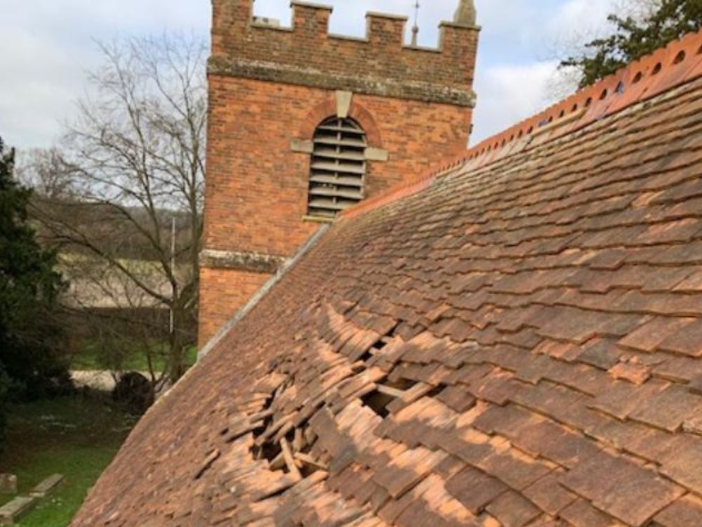 Close-up view of a red tile church roof in poor condition with several broken and slipped tiles opening up holes in the roof. A crenellated red brick church tower can be seen in the background. (c) Peter Hawkins