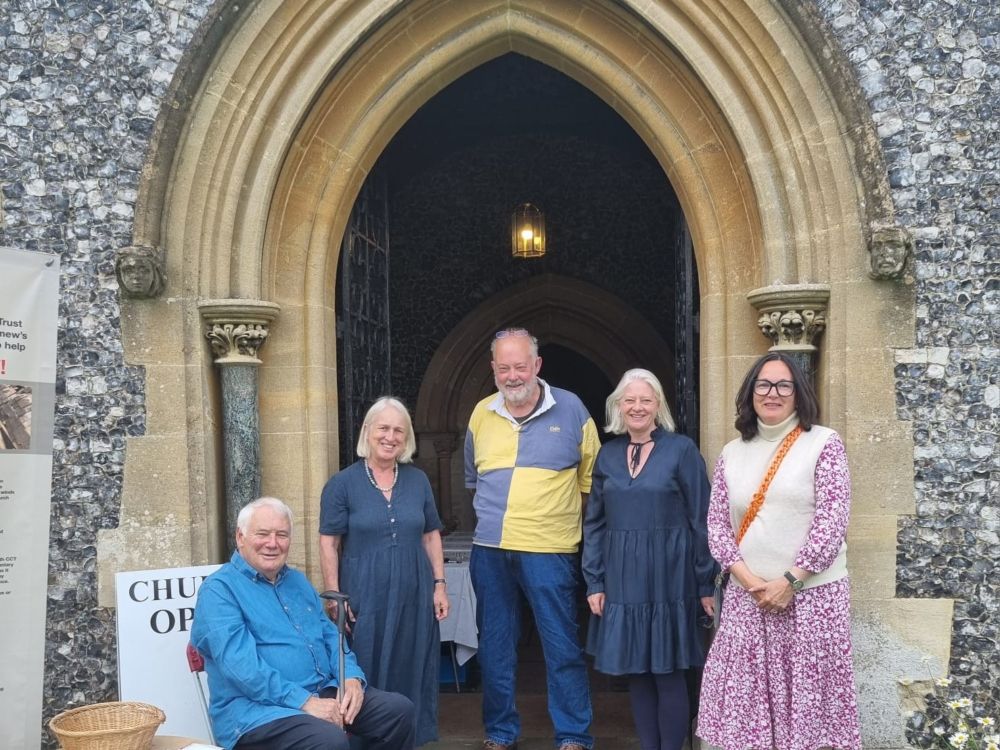 Five people posing for a photograph in front of a pointed church archway. All are smiling, a man on the left in blue shirt is sitting down, to his right stands a woman in blue dress, next to her a tall man in jeans and blue and yellow checkered jumper, to his right another woman in a blue dress almost identical to the first woman's dress and to her right stands a woman in a pink dress, with a white vest top. All have grey hair, except the woman on the far right, who has black hair.