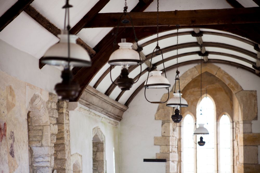 Interior of Whitcombe church, a small single cell building, with whitewashed, partly exposed walls, a three light east or west window and dark wooden roof beams. Suspended from the ceiling are several white gas lamps in a neat row. (c) Andy Marshall