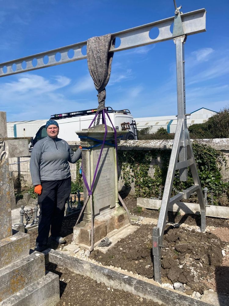 A member of the group standing next to a gravestone being suspended from a beam, wearing orange gloves.
