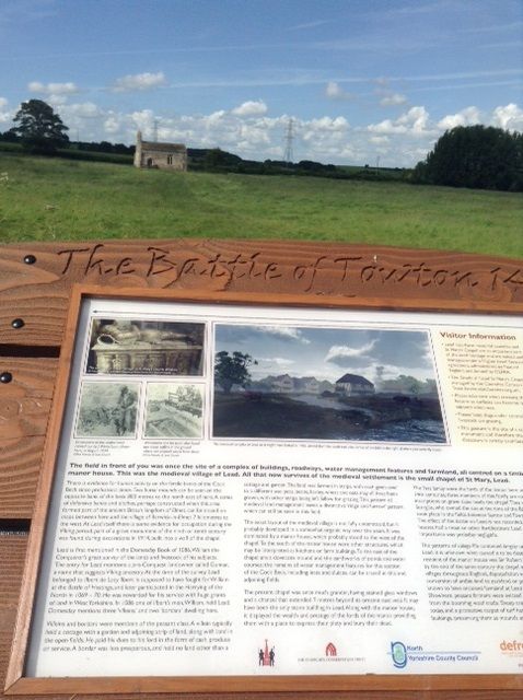 A photograph of a sign set in a wooden frame engraved with text saying 'The Battle of Towton' with images and text relating to the battle underneath. In the background we can see St. Mary's lead under bright blue sky. 