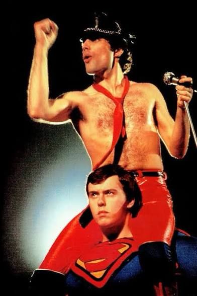 Freddie Mercury shirtless in a black leather cap, red vinyl(?) pants and a red tie on the shoulders of another Superman who is taking his job very seriously. 