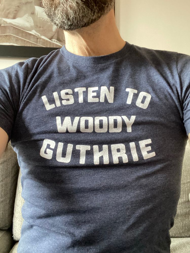 A dark heather gray t-shirt that says “LISTEN TO WOODY GUTHRIE” in all-caps. 