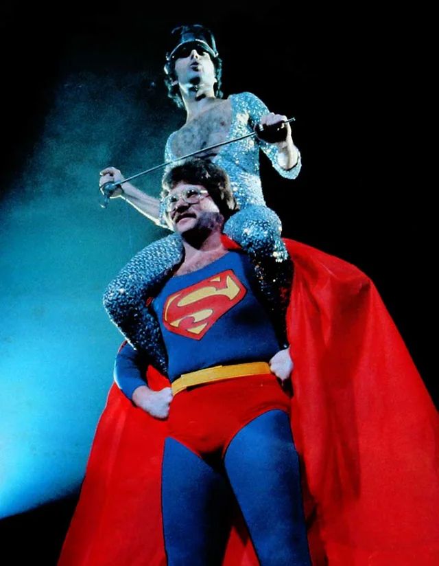 Freddie Mercury in a glittery leotard on the shoulders of a Superman wearing fancy glasses