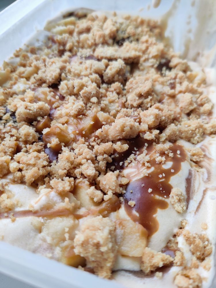 A tub of ice cream with pieces of apple and crumble sat on the top and swirls of dark caramel sauce.