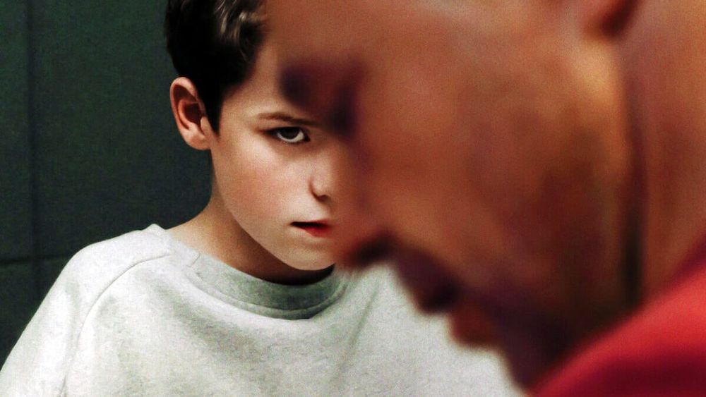 Le père au premier plan flou détournant son regard sur sa gauche, son tshirt rouge couvrant son épaule gauche est visible en bas à droite. Le focus est à l'arrière plan sur le fils en survèt gris, cheveux noirs, le regard fixé sur son père, on voit la moitié droite de son visage. 