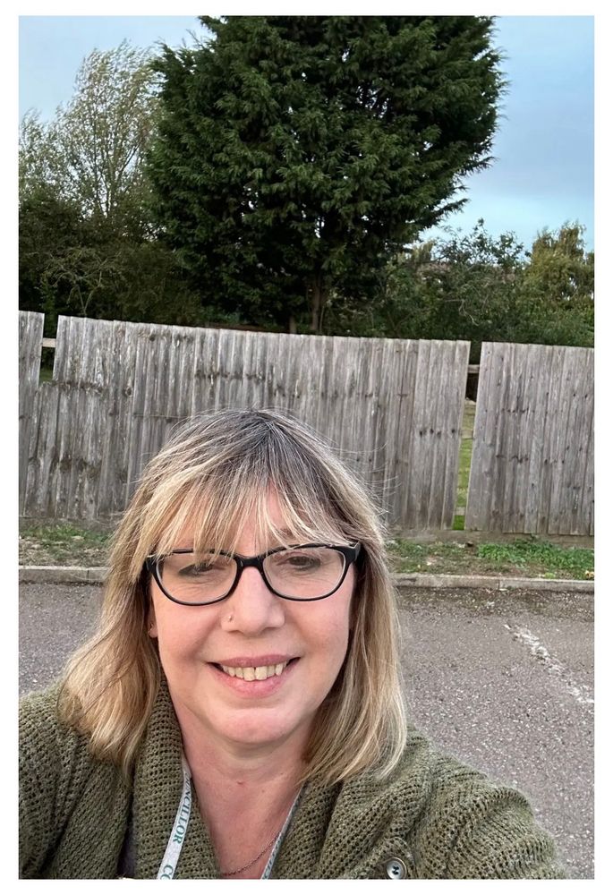 Nicola takes a selfie stood in front of a wooden fence and a tall tree, she’s a white lady with shoulder length blonde hair with brown undertones, she’s wearing black framed glasses and is smiling at the camera, she’s wearing a white and green lanyard around her neck.