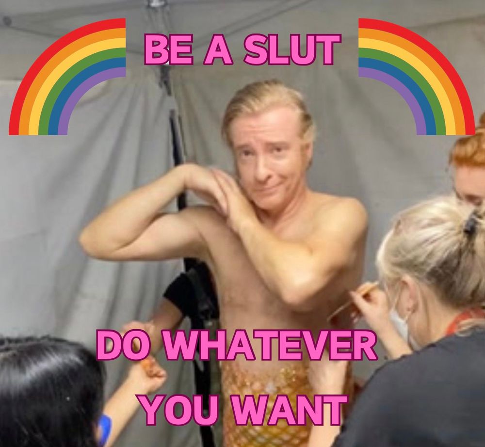 Rhys Darby in bts getting dressed up as a mermaid and the text says “be a slut do whatever you want” with rainbows