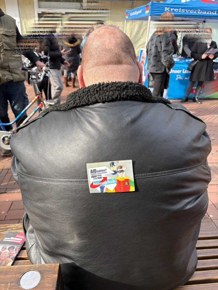 Glatzköpfiger Mann mit Lederjacke, auf einer Bank sitzend. Auf seinem Rücken klebt ein Aufkleber. Der Aufkleber zeigt einen Comic-Helden (angelehnt an Robin von Batman) und trägt folgenden Text: „AfD wählen?  kannste schon machen, aber dann bist du halt homophob“. Im Hintergrund sind mehrere Menschen vor einem AfD-Stand mit einem blauen Pavillon zu sehen. Auf dem Pavillon steht „Kreisverband“. Auf der Bank neben dem Mann liegen noch weitere Flyer. Die Szene spielt sich augenscheinlich bei einer Kundgebung oder Veranstaltung im Freien ab.