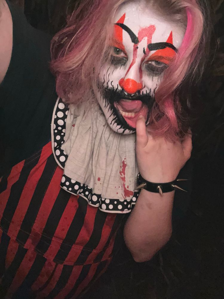 a clown with orange and black corpse make up in orange and black overalls staring into the camera with a white collar with bubble patterned fringe