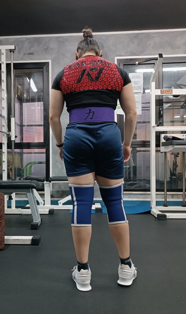 Person in a gym seen from the back, with specialized attire on. Supportive knee sleeves, squat shoes, bar grip shirt, lever belt for powerlifting.