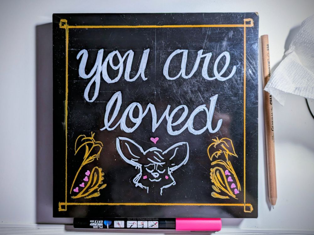 a black square panel with "you are loved" painted on in white script text. below is the simplified bust of a big-eared fox, eyes closed contentedly and blushing, with a heart overhead. It's all bordered in gold with flowery flourishes