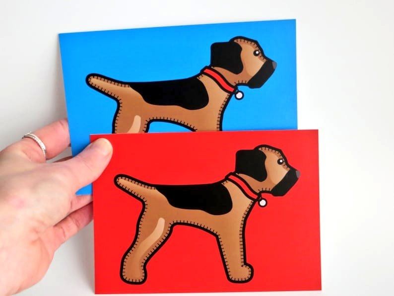 A hand holding a red and a blue A6 postcard print of the misheleneous border terrier logo dog