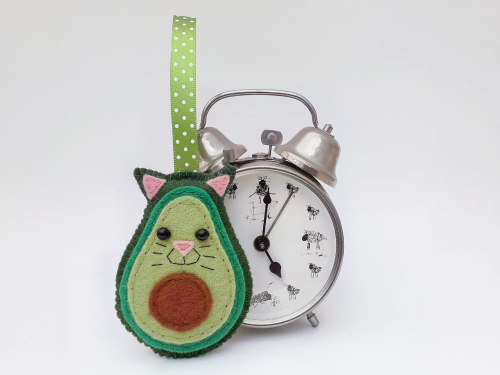 A handmade felt kawaii avo-cat-o ornament with feline features and ears by misheleneous. Displayed against a retro silver alarm clock
