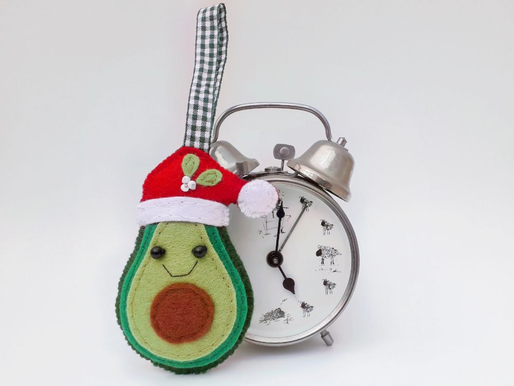 A handmade felt kawaii avocado-ho-ho ornament by misheleneous wearing a red santa hat with mistletoe detail. Displayed against a retro silver alarm clock