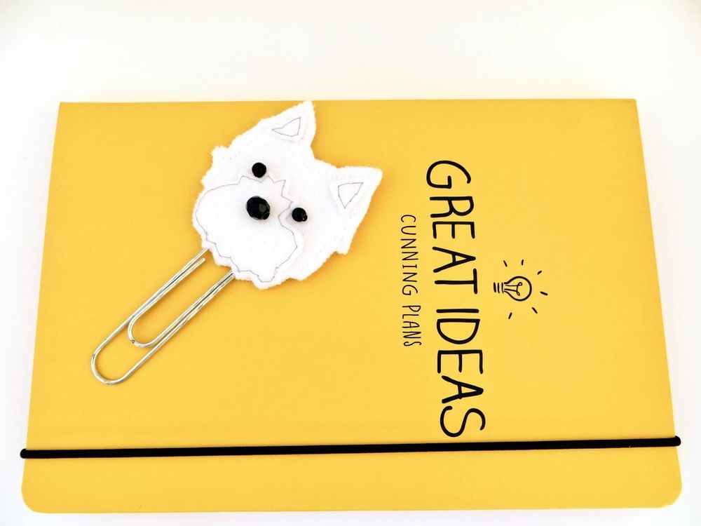 A handmade felt planner clip bookmark by misheleneous featuring a white west highland terrier dog head topper. Displayed on a yellow journal