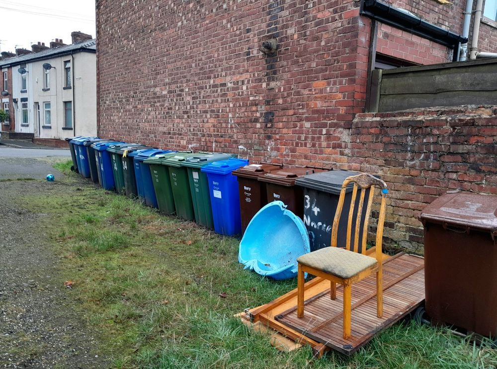 A row of wheely bins along a terrace gable end wall with a random dining chair and a blue plastic sandpit or paddling pool. Prestwich, Manchester