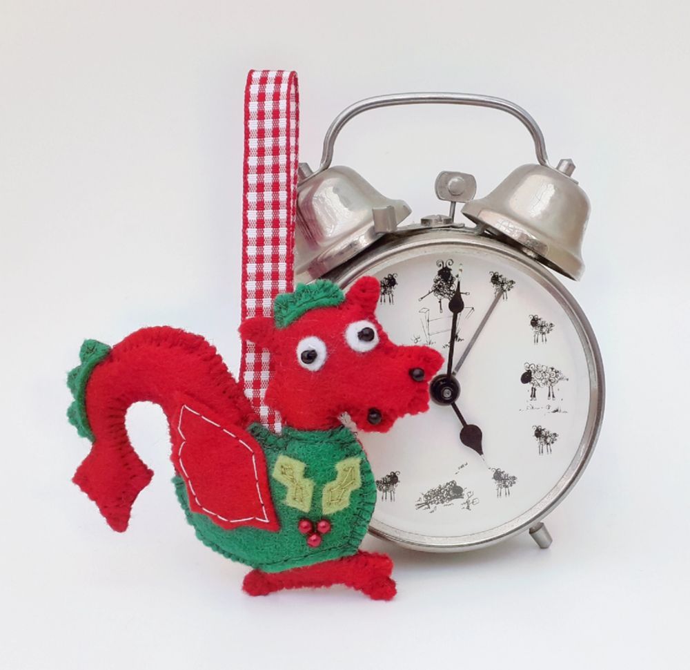 A handmade felt red welsh dragon christmas ornament by misheleneous wearing a green festive jumper with holly motif. Displayed next to a silver retro alarm clock