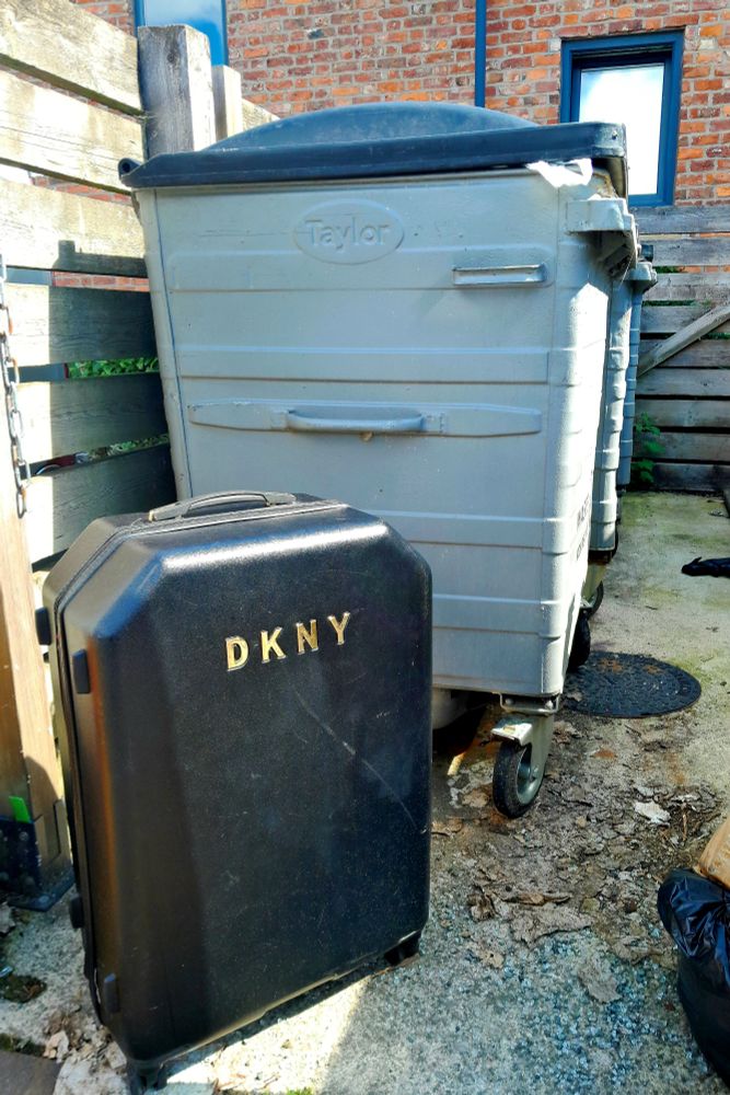 A black moulded DKNY suitcase abandoned next to big grey wheely bins in Prestwich, Manchester