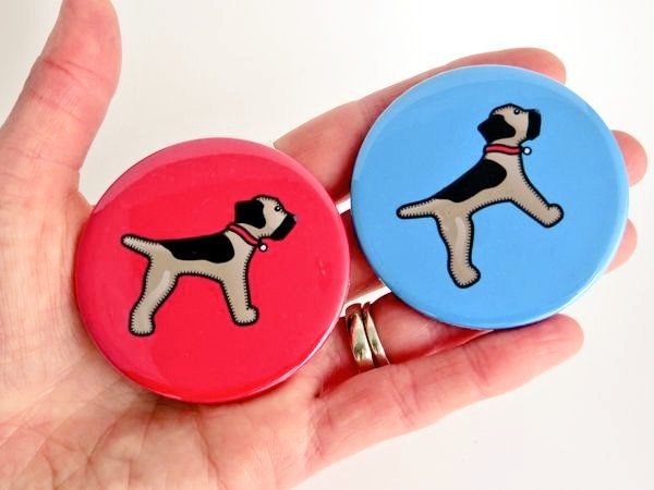 A hand holding a red and a blue pocket mirror printed with the misheleneous border terrier logo dog